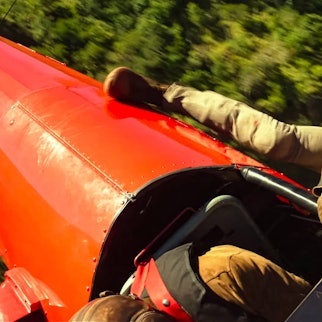Tom Cruise hängt als Ethan Hunt an einem fliegenden, roten Doppeldecker-Flugzeug.