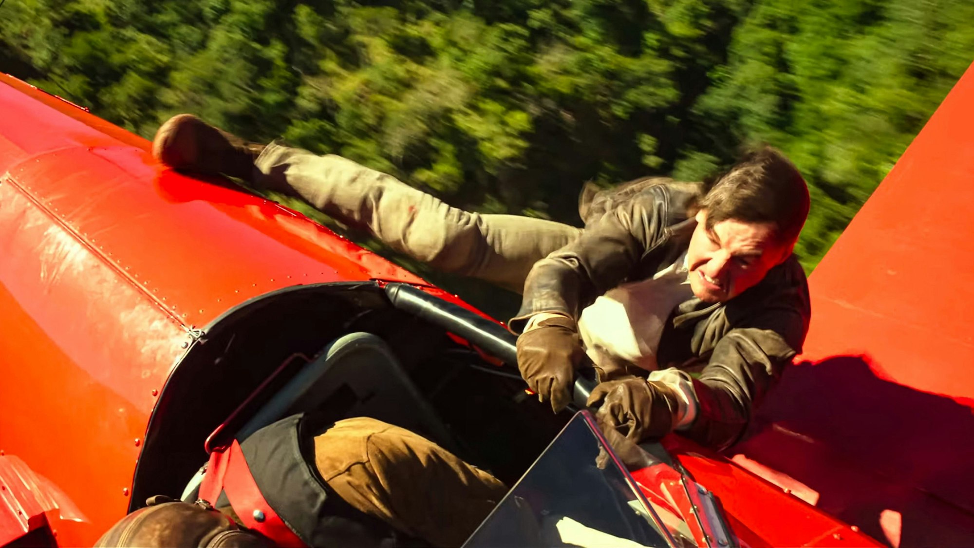 Tom Cruise hängt als Ethan Hunt an einem fliegenden, roten Doppeldecker-Flugzeug.