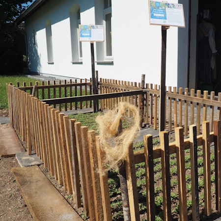 Ein Zaun steht um ein kleines Beet, in dem ein Schild steckt. An einem Zaunpfahl hängt ein Büschel Flachs-Fasern.