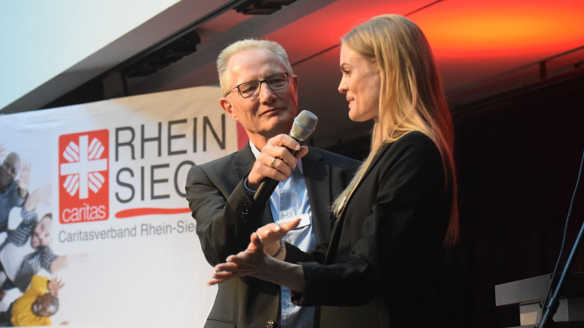 Ein Mann und eine Frau auf einer Veranstaltungsbühne. Im Hintergrund steht eine Werbetafel mit der Aufschrift Caritasverband Rhein-Sieg.