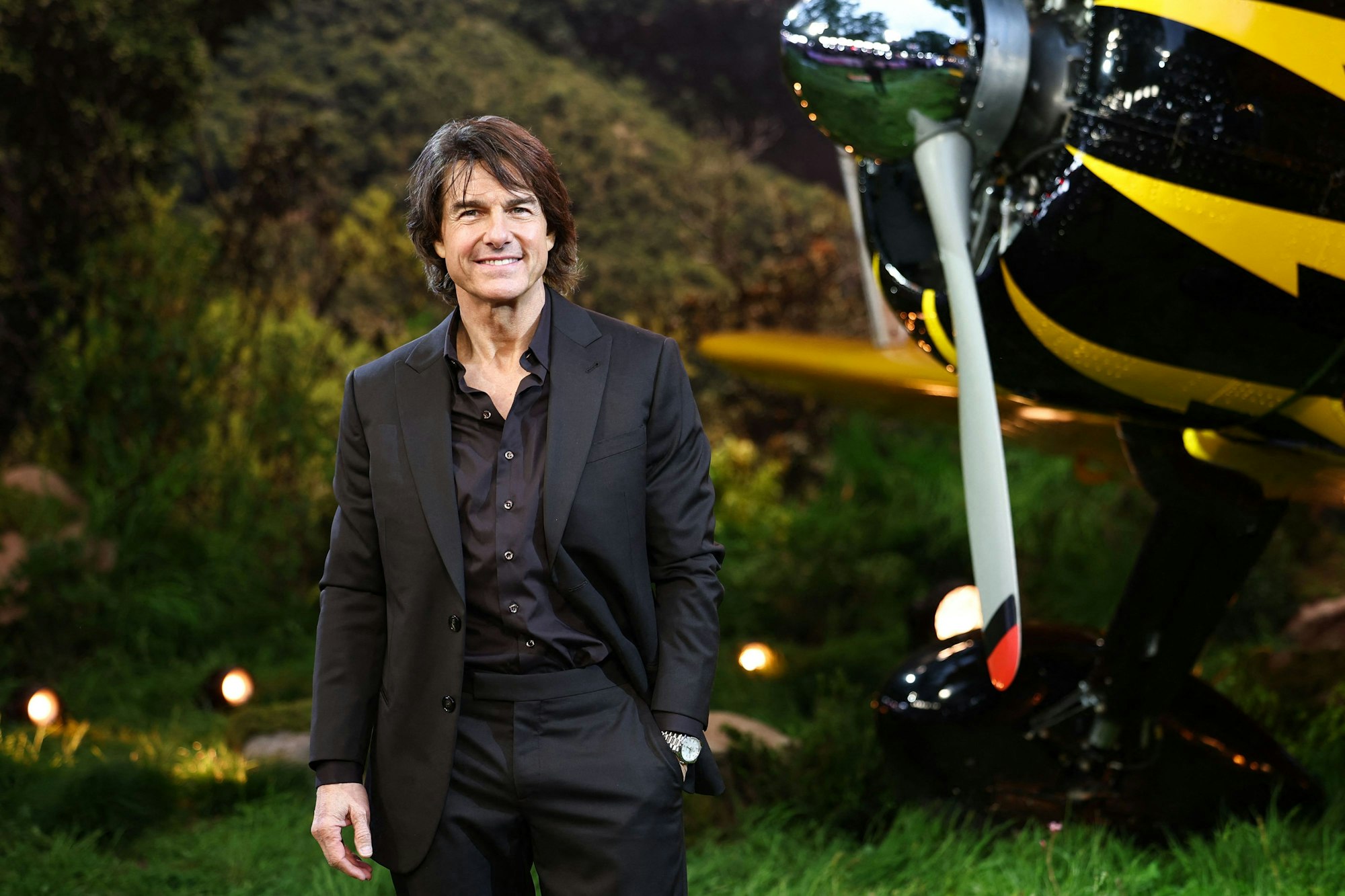 Tom Cruise steht im schwarzen Anzug vor einem gelben Flugzeug.