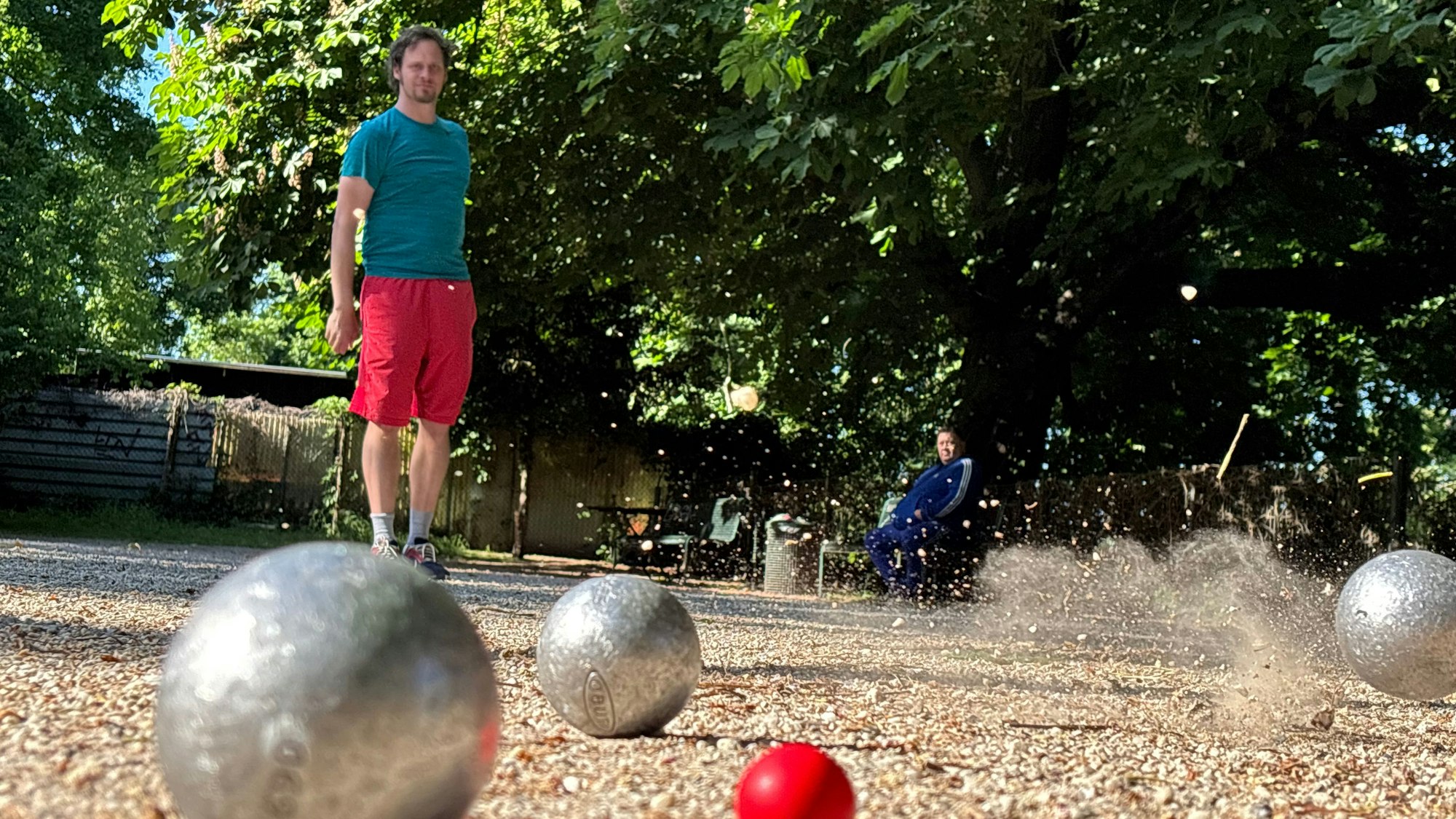 Ein Mann in sportlicher Hose und T-Shirt hat eine Boule-Kugel geworfen, die beim Aufprall Sand und Steinchen in die Luft schleudert.