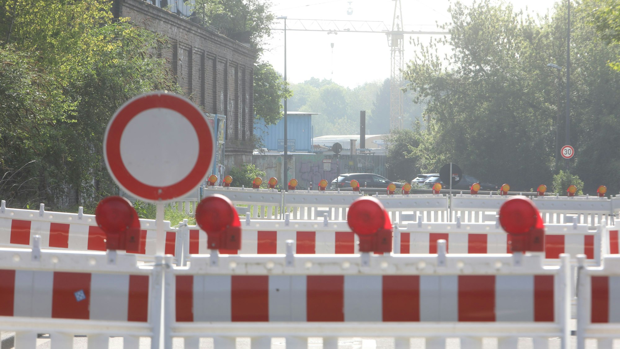 Rot-weiße Absperrbaken stehen auf einer Straße, an deren Rand alte Industrie-Backsteingebäude stehen.
