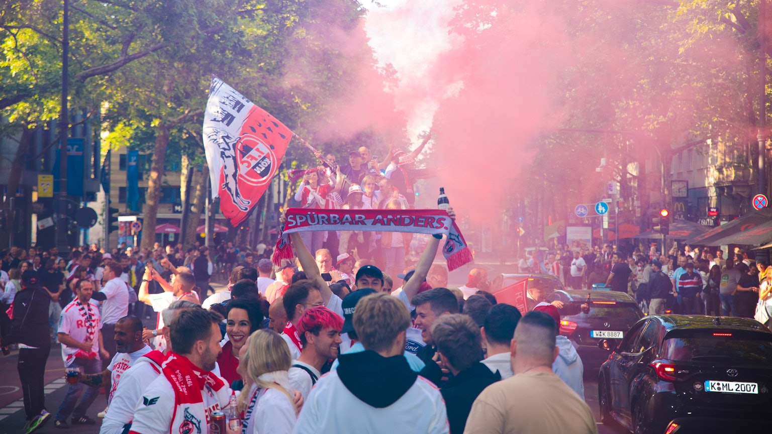Menschen mit Fanschals des 1. FC Köln knubbeln sich auf einer Straße.