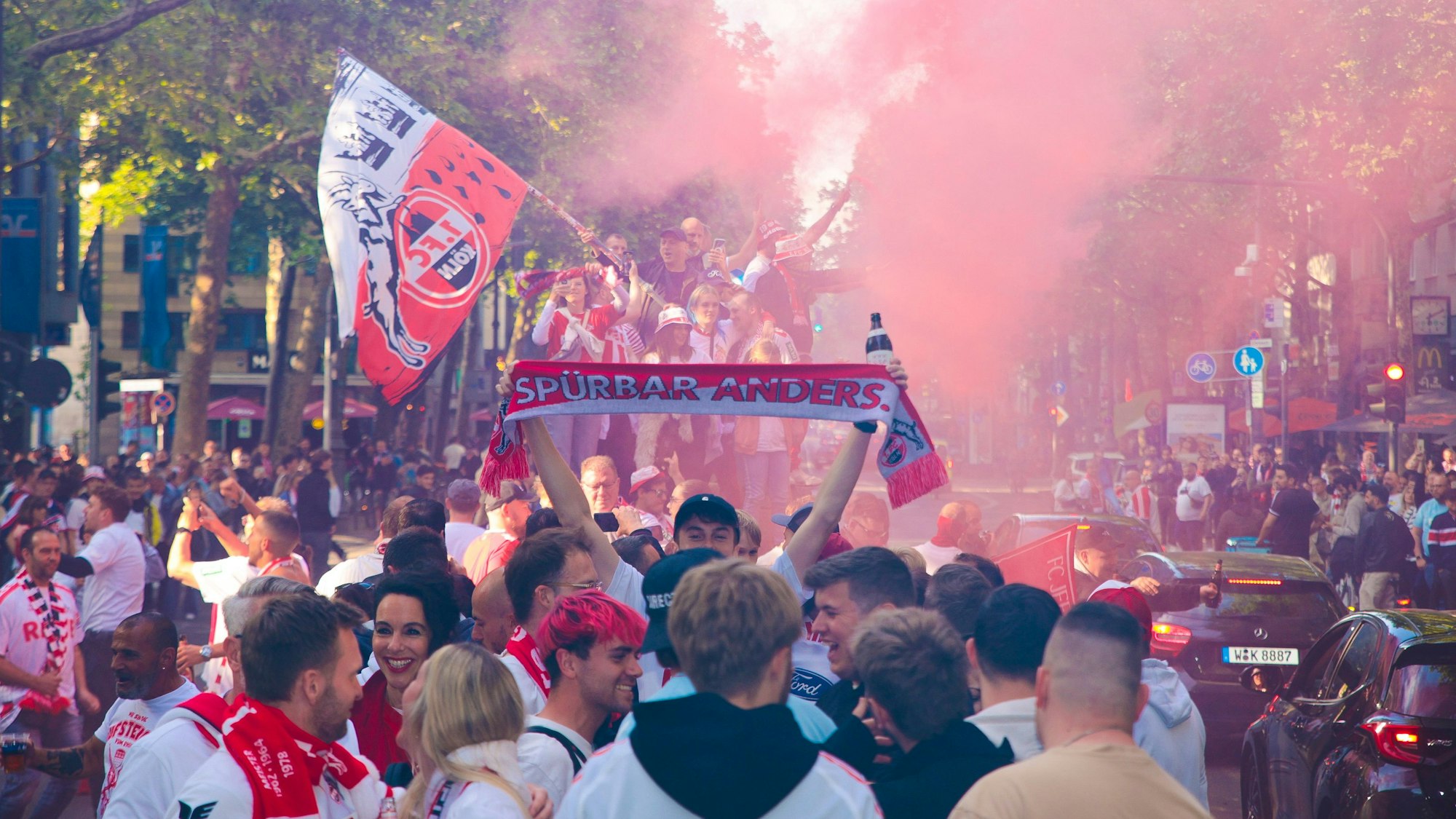 Menschen mit Fanschals des 1. FC Köln knubbeln sich auf einer Straße.