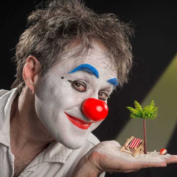 Clown Calvero ist geschminkt und hält eine Palme und einen Liegestuhl im Miniformat auf der Hand.