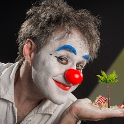 Clown Calvero ist geschminkt und hält eine Palme und einen Liegestuhl im Miniformat auf der Hand.