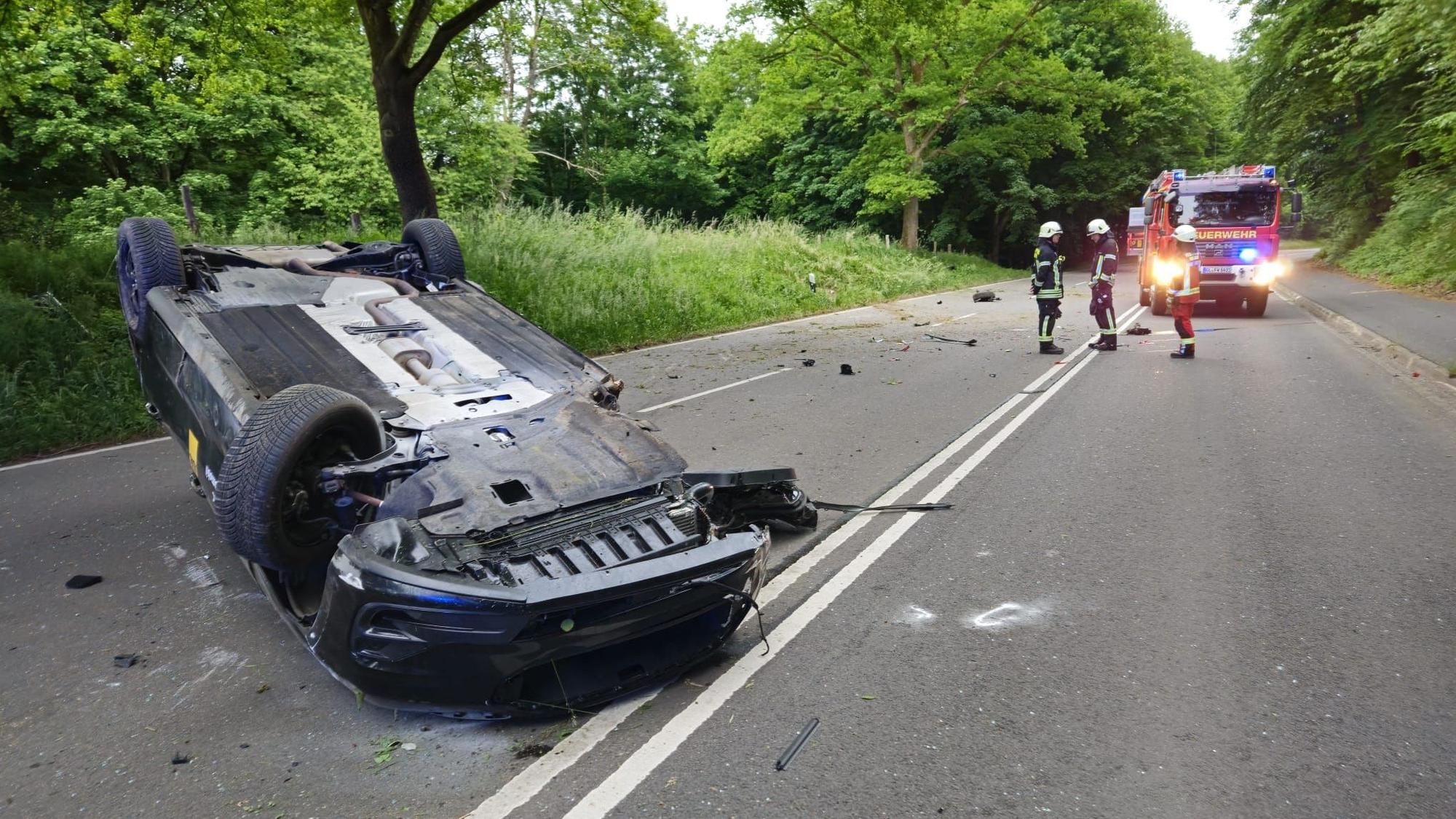 Ein Auto liegt auf dem Dach am Heiligenhauser Berg in Overath. Im Hintergrund stehen Einsatzkräfte und ein Feuerwehrfahrzeug.