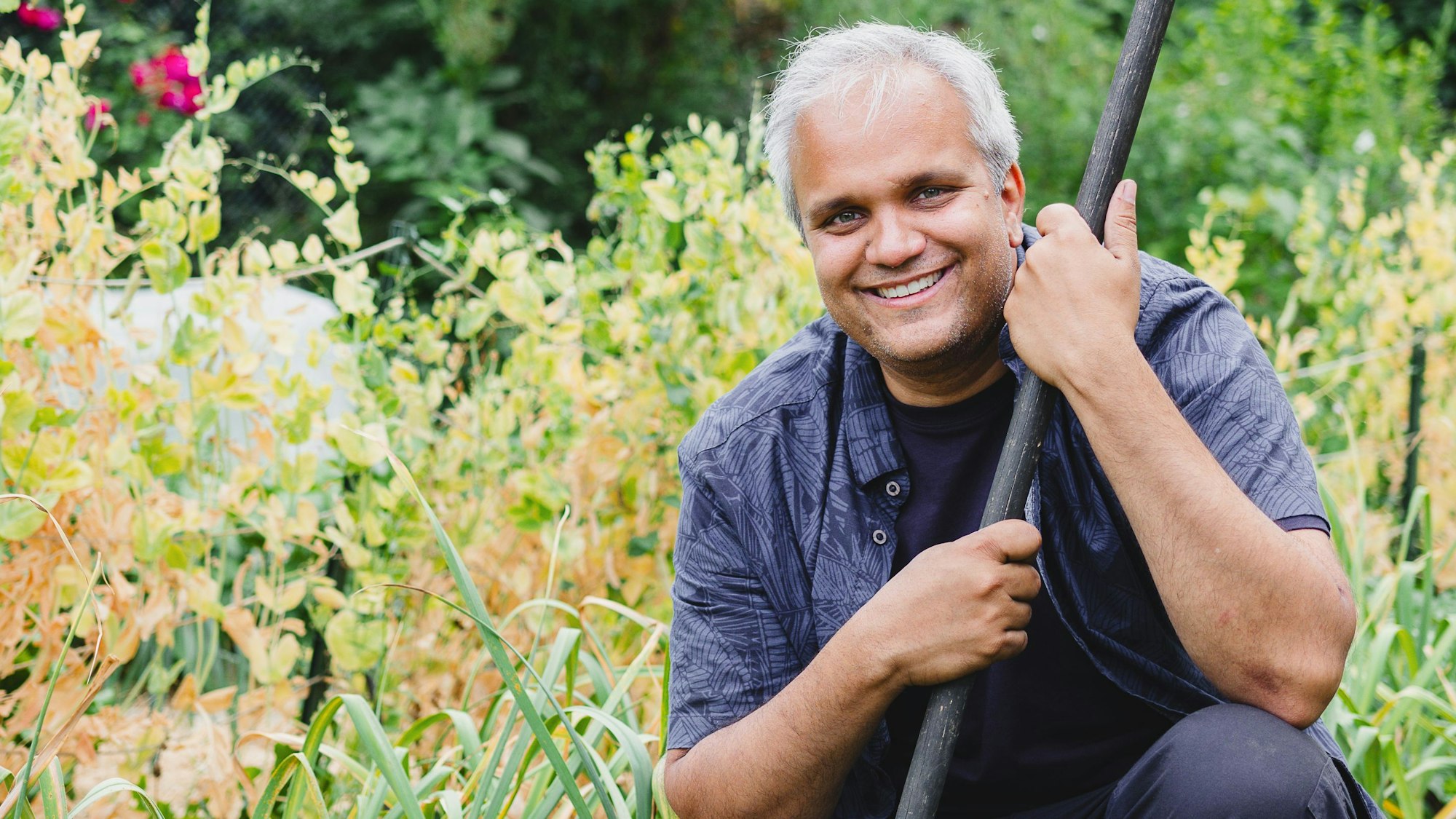 Auf dem Bild ist ein Mann zu sehen, der in seinem Garten auf der Erde hockt.
