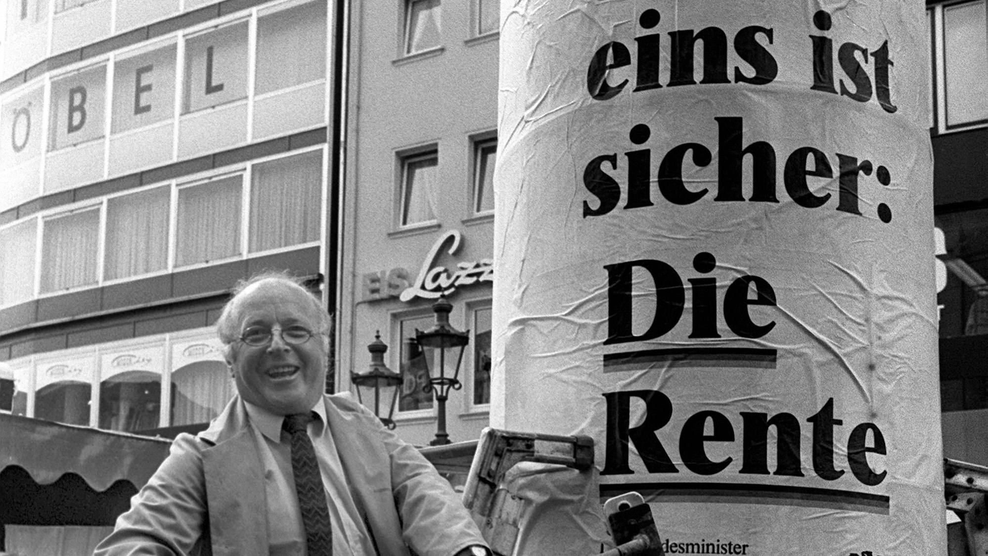 Auf einem Schwarzweiß-Foto ist der ehemalige Bundesarbeitsminister Norbert Blüm (CDU) im Arbeitskittel zu sehen, der eine Litfaßsäule mit dem Plakat „Denn eins ist sicher: Die Rente“ beklebt. Das Foto stammt aus dem Jahr 1986.