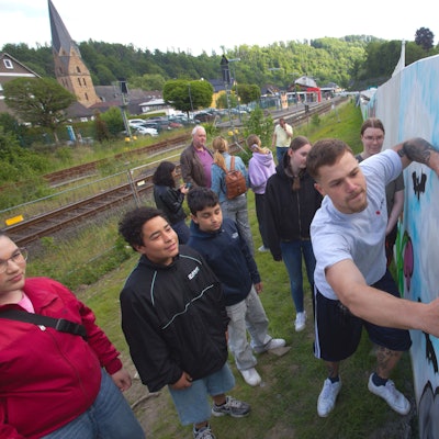Das Foto zeigt einen Sprayer an einer Mauer und jugendliche Zuschauer.