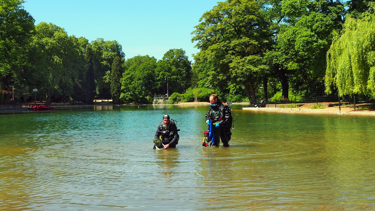 Zwei Männer in Taucheranzügen stehen in einem Weiher. Einer hat eine Pflanzkiste in der Hand.