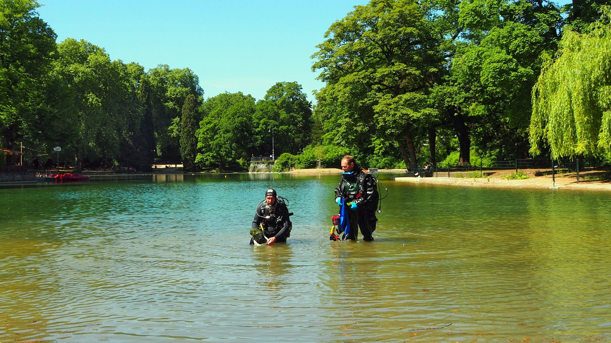 Zwei Männer in Taucheranzügen stehen in einem Weiher. Einer hat eine Pflanzkiste in der Hand.