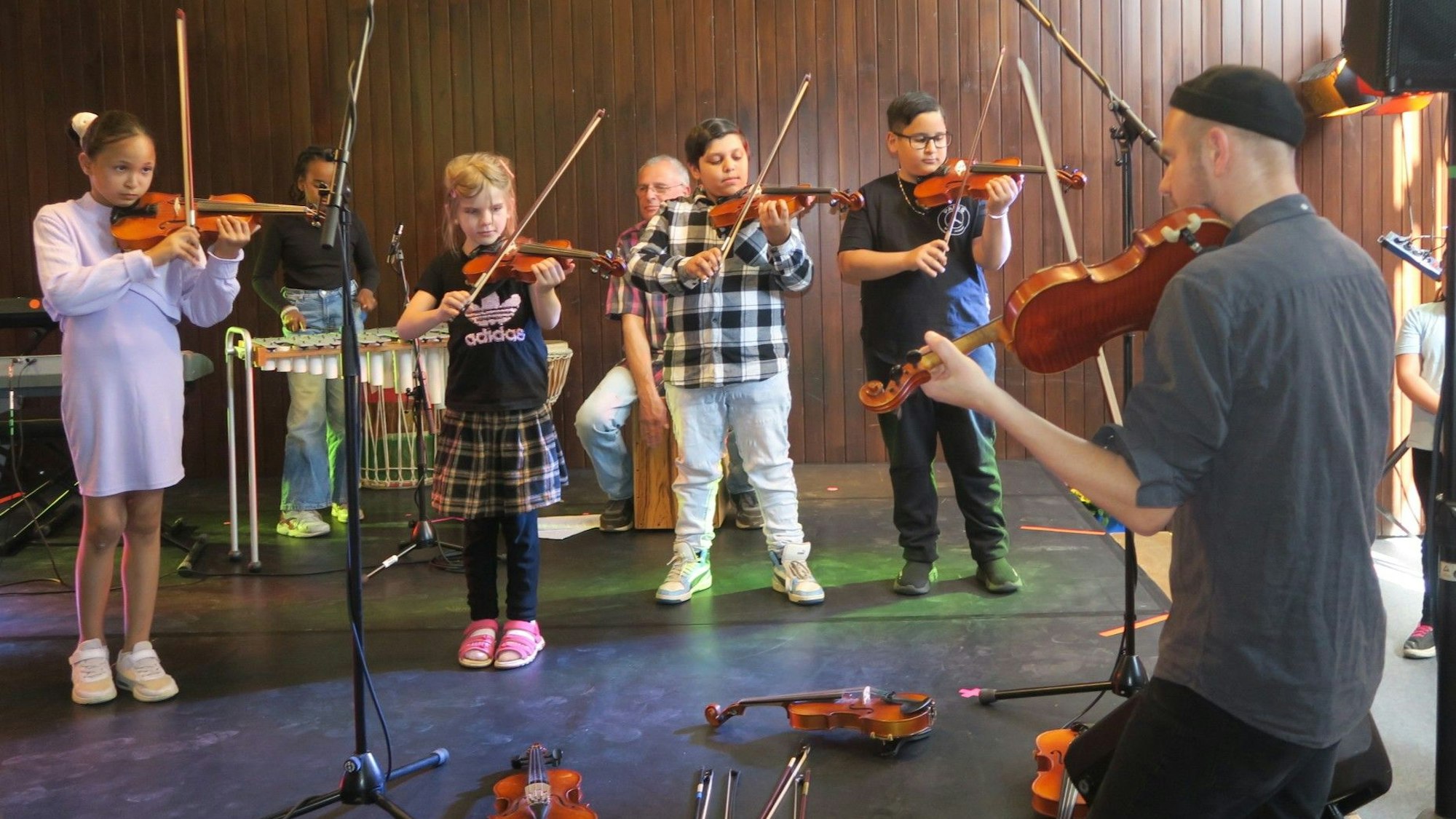 Einige Kinder stehen auf einer Bühne, mit dem Rücken zur Kamera steht ein Mann mit Geige.