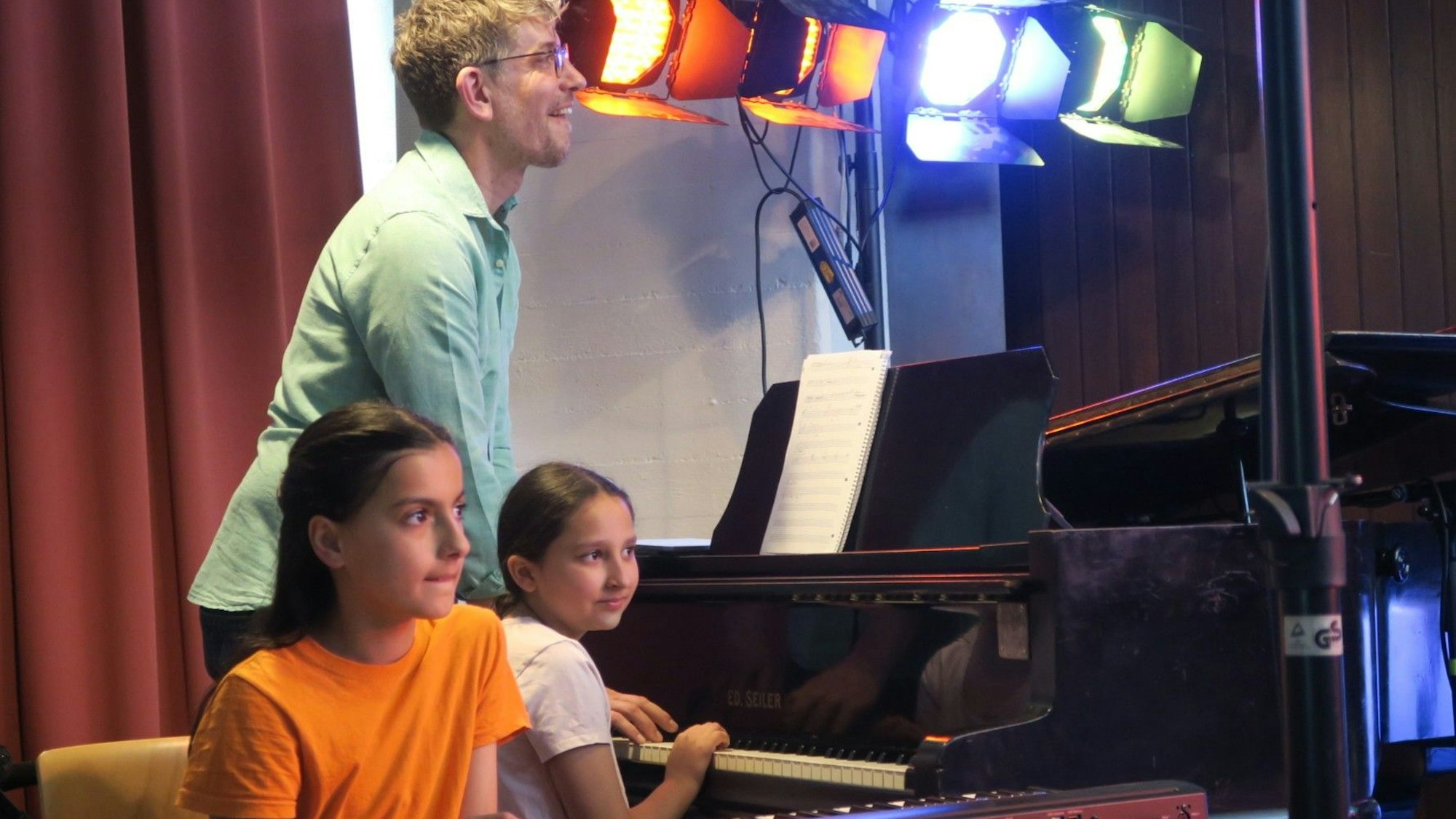 Ein Mann steht am Klavier, zwei Mädchen sitzen an Keyboard und Piano.