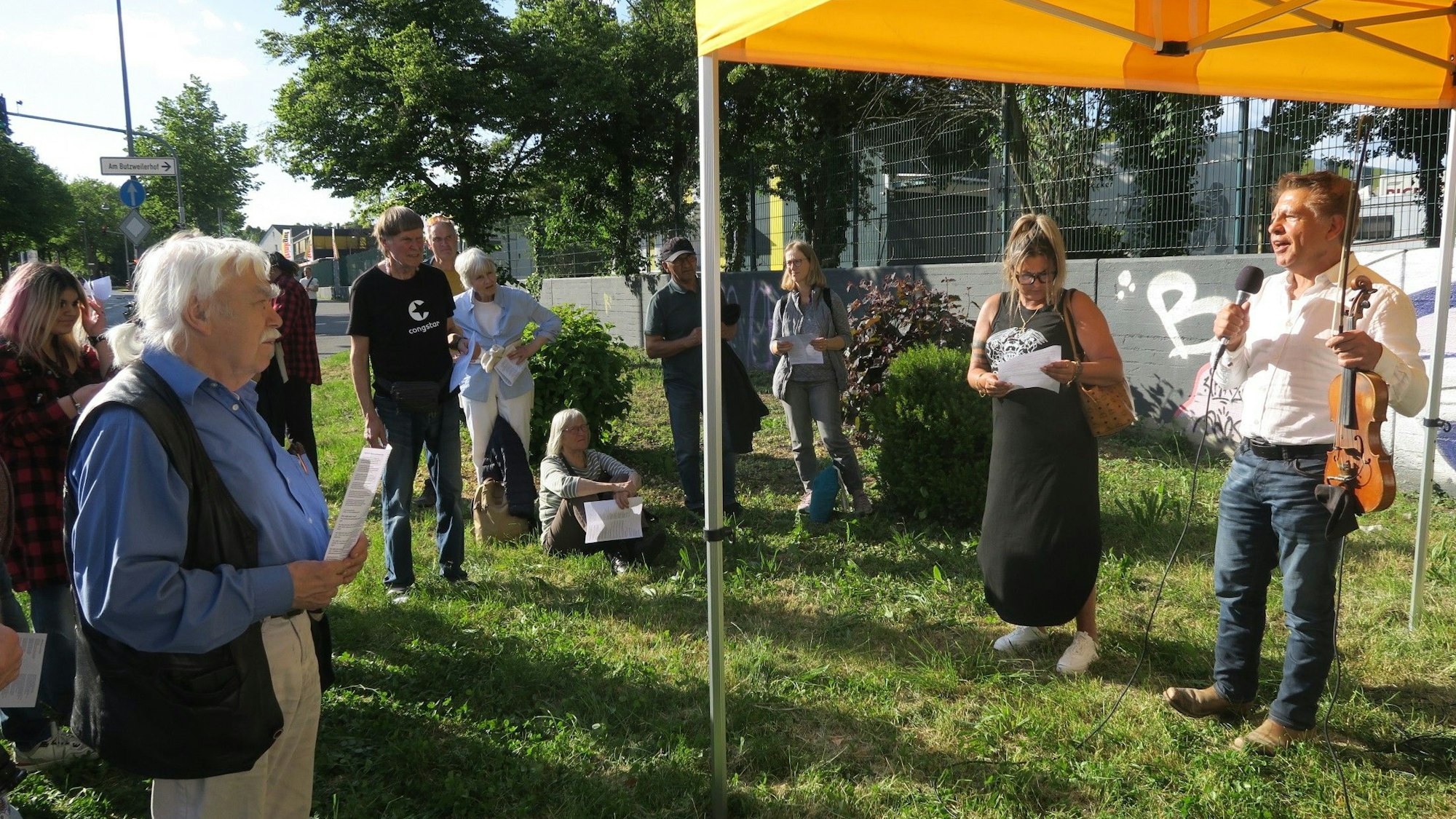 Menschen stehen auf einer Wiese. Ein Mann spricht in ein Mikrofon und hält eine Geige in der Hand.