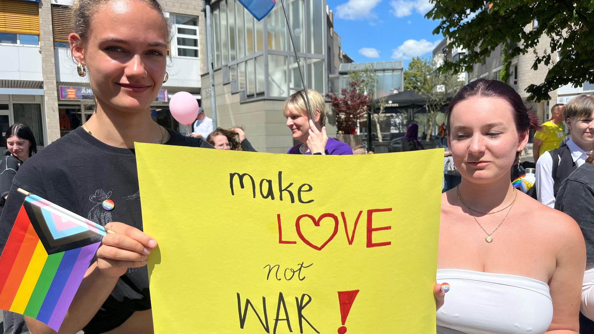 „Make Love not War“ steht auf einem gelben Plakat, dass zwei junge Frauen zwischen sich halten.