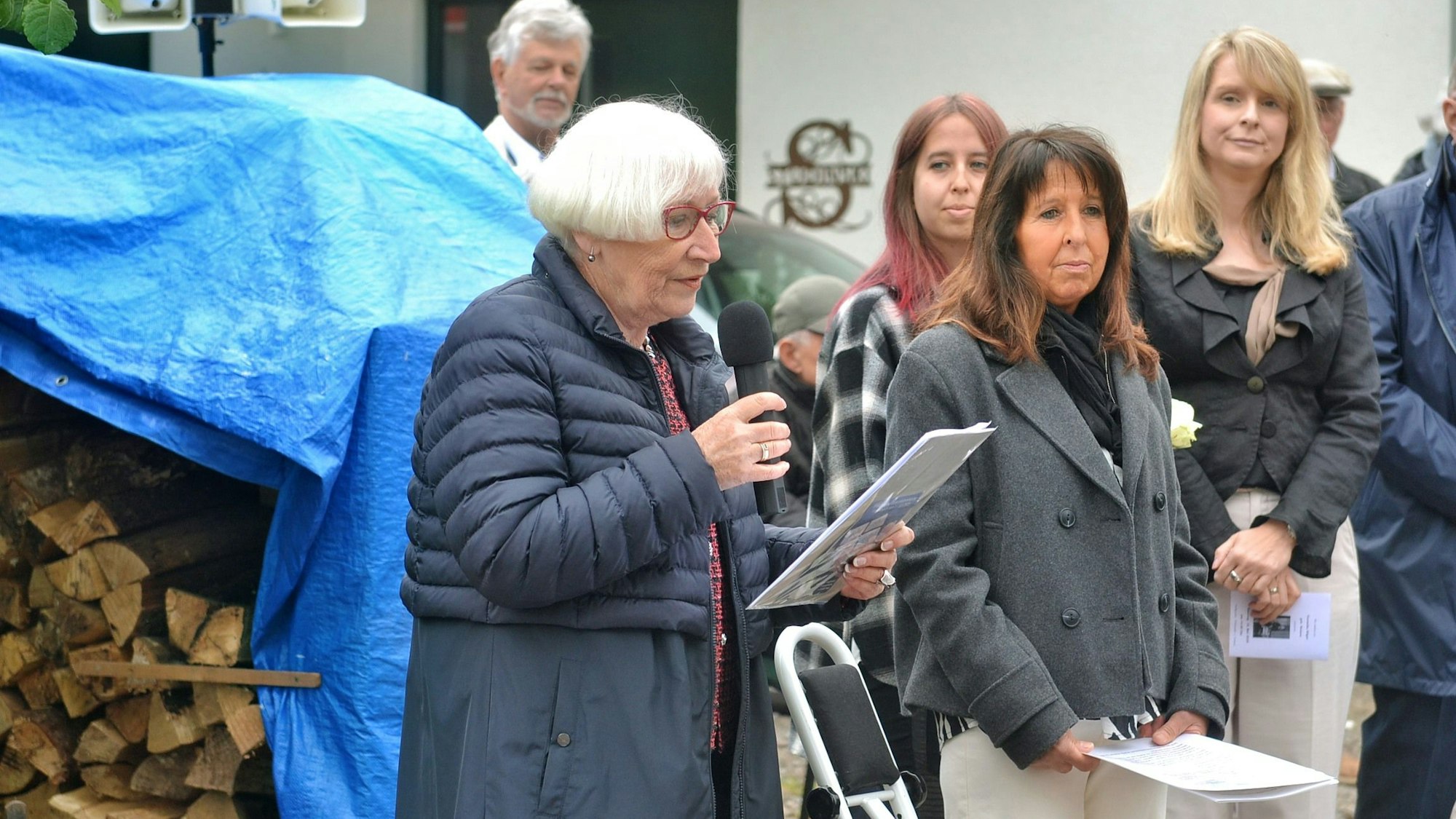 Gisela Freier hält ein Mikrofon und ein Manuskript in den Händen. Daneben stehen einige der Besucher.