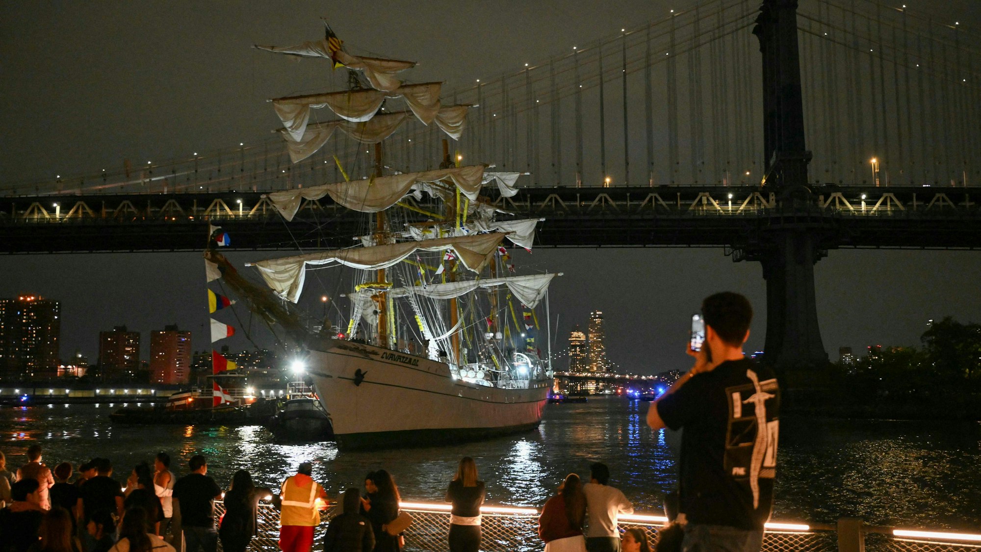 People watch as a Mexican Navy training ship is pulled away after it slammed into the nearby Brooklyn Bridge in New York on May 17, 2025. A Mexican Navy training ship slammed into the Brooklyn Bridge late Saturday, snapping all three of its masts and igniting a rescue operation beneath the iconic New York City landmark. The Mexican Navy said in a statement that 22 people on board the training ship were injured, three of them critically. (Photo by ANGELA WEISS / AFP)