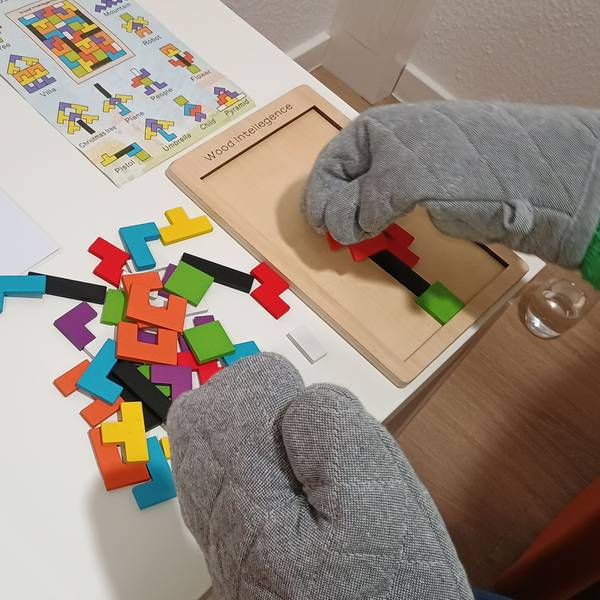 Das Bild zeigt eine Mitarbeiterin, die mit Ofenhandschuhen ein Holzpuzzle macht. Die Ofenhandschuhe simulieren eine taktile Unterempfindlichkeit und erschweren eine feinmotorische Aufgabe. Viele Menschen mit Autismus haben solche Wahrnehmungsbesonderheiten, auch wenn sie nicht zur Kernsymptomatik gehören.