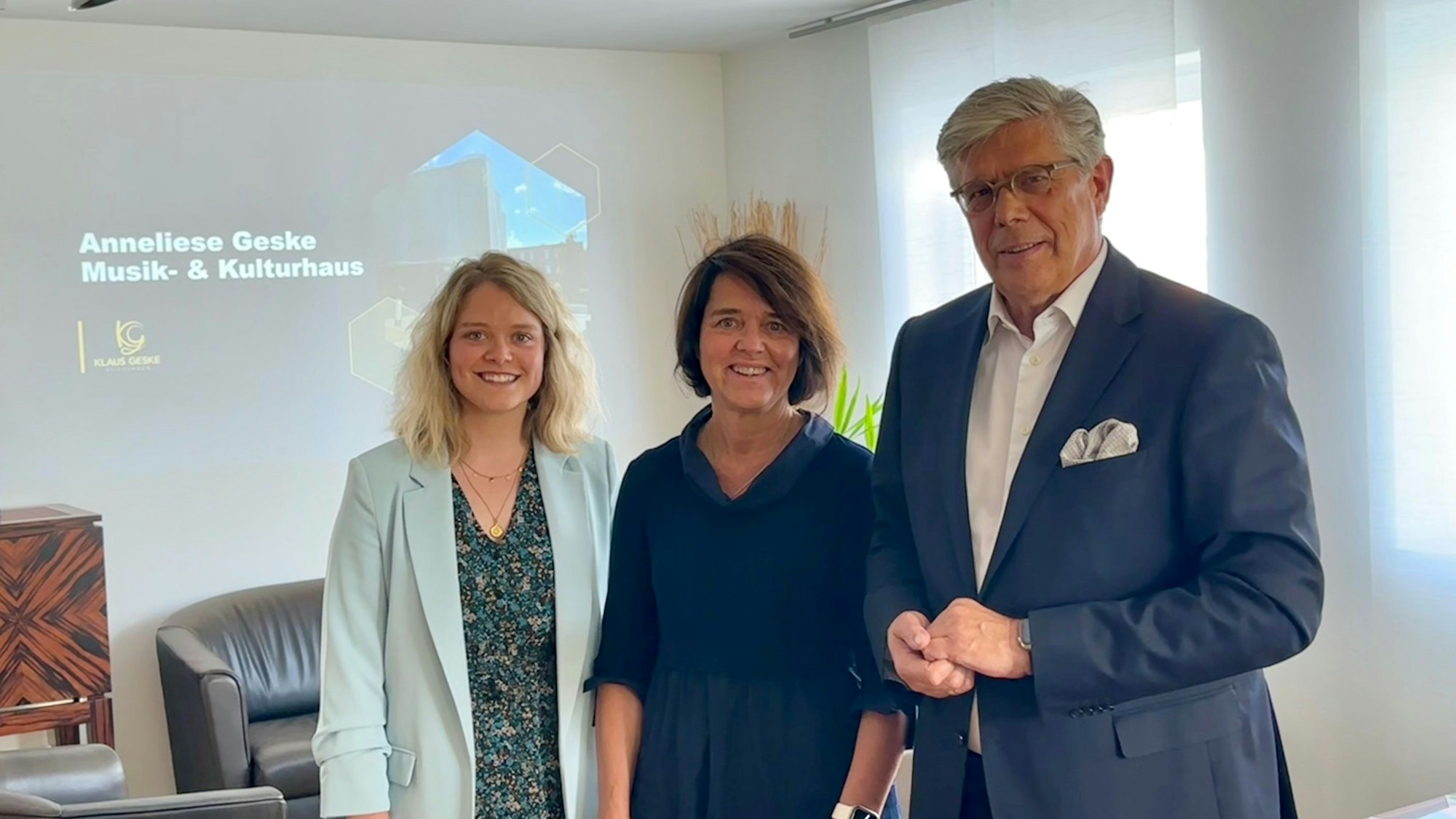 Zwei Frauen und ein Mann stehen vor einem Hintergrund, auf dem „Anneliese Geske Musik- und Kulturhaus“ steht.