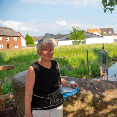 Anita Scholl steht an einer Steinbrüstung, hinter ihr sind ein Bachlauf ein Wehr, eine Wiese und Häuser zu sehen.