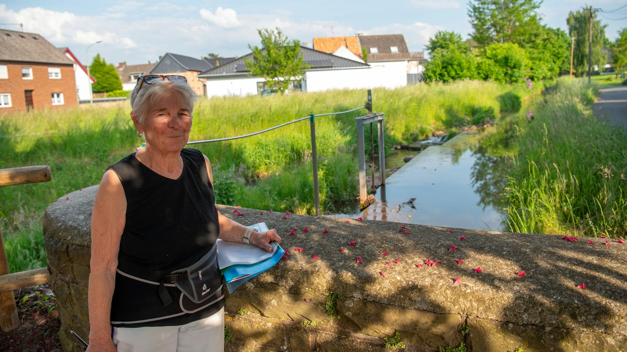 Anita Scholl steht an einer Steinbrüstung, hinter ihr sind ein Bachlauf ein Wehr, eine Wiese und Häuser zu sehen.