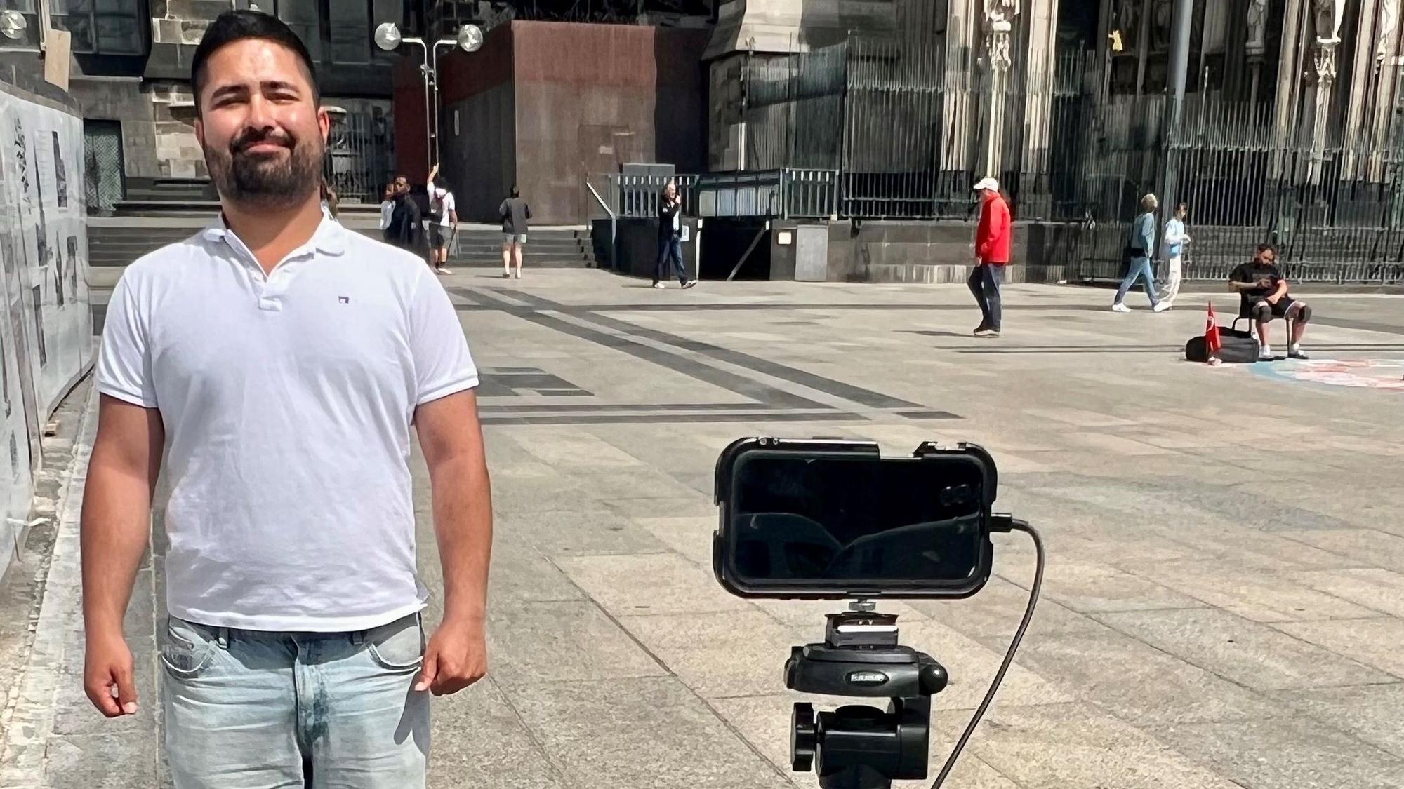 Zabur Asadi steht vor dem Kölner Dom. Vor ihm ist ein Handy als Kamera aufgestellt.