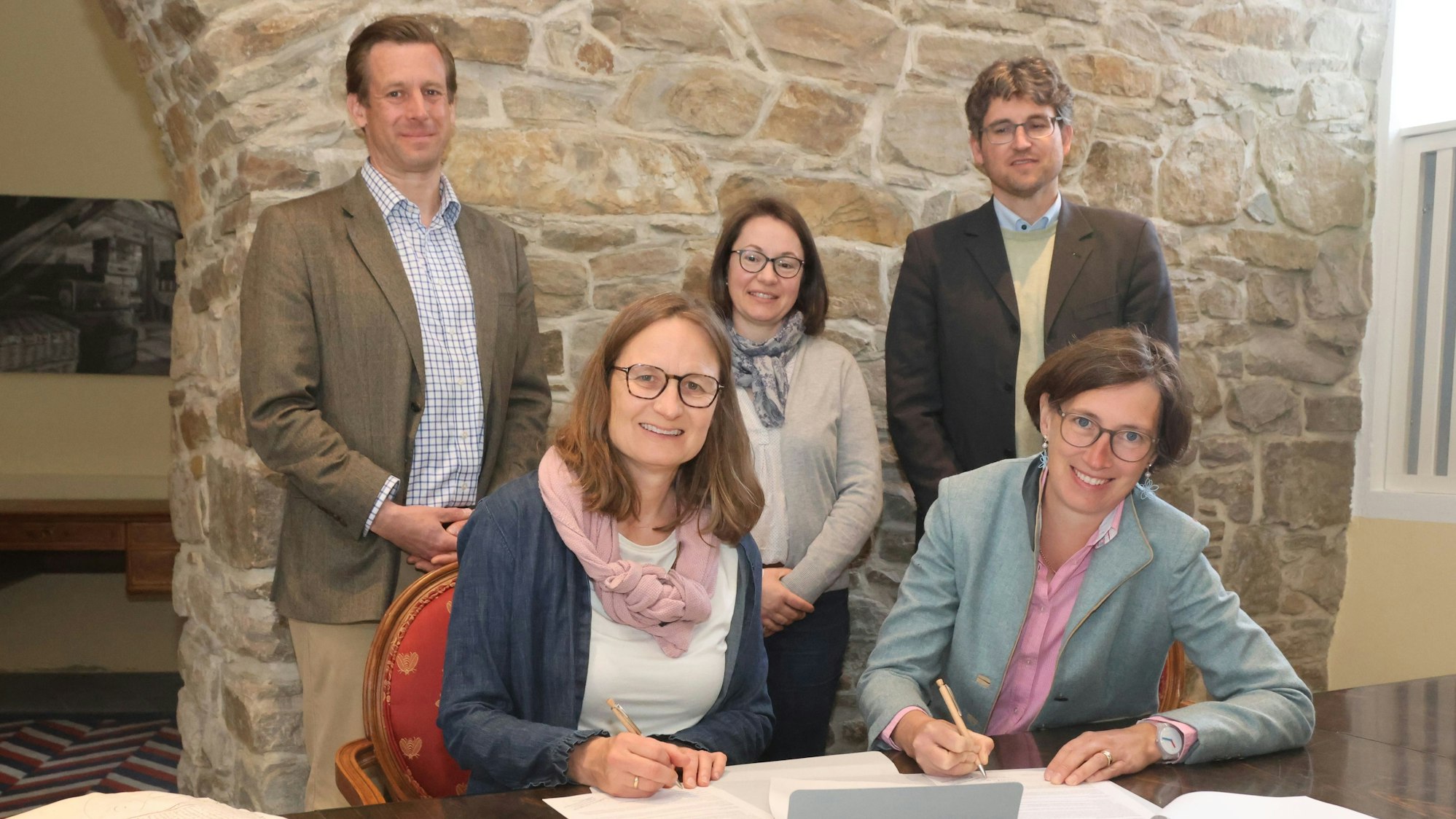 Baronin Elisabeth von Lüninck (r.) und Angela Heienbrok sitzen an einem Tisch und unterzeichnen die Vereinbarung, dahinter Clemens Freiherr von Boeselager (v.l.), Heike Bartel-Heuwinkel und Gregor Patt.