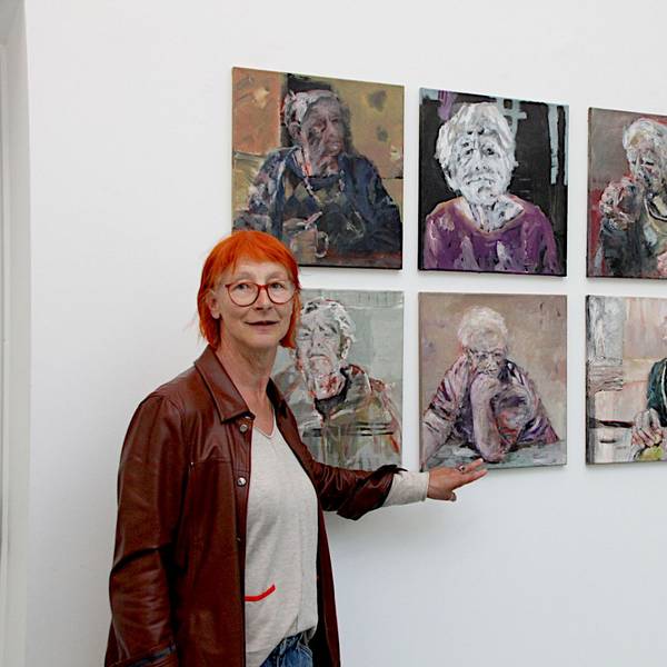 Die Künstlerin Eva Görgen, die rote Haare und eine Lederjacke trägt, steht vor neun ihrer Werke, die jeweils ein Porträt einer älteren Frau zeigen.