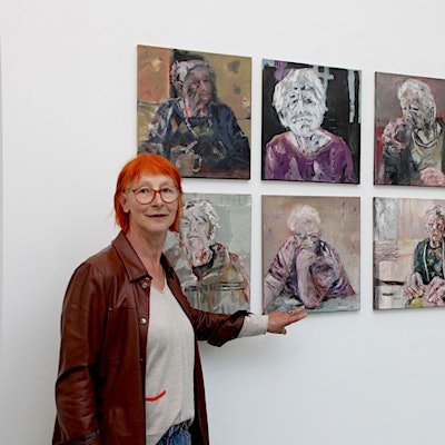 Die Künstlerin Eva Görgen, die rote Haare und eine Lederjacke trägt, steht vor neun ihrer Werke, die jeweils ein Porträt einer älteren Frau zeigen.