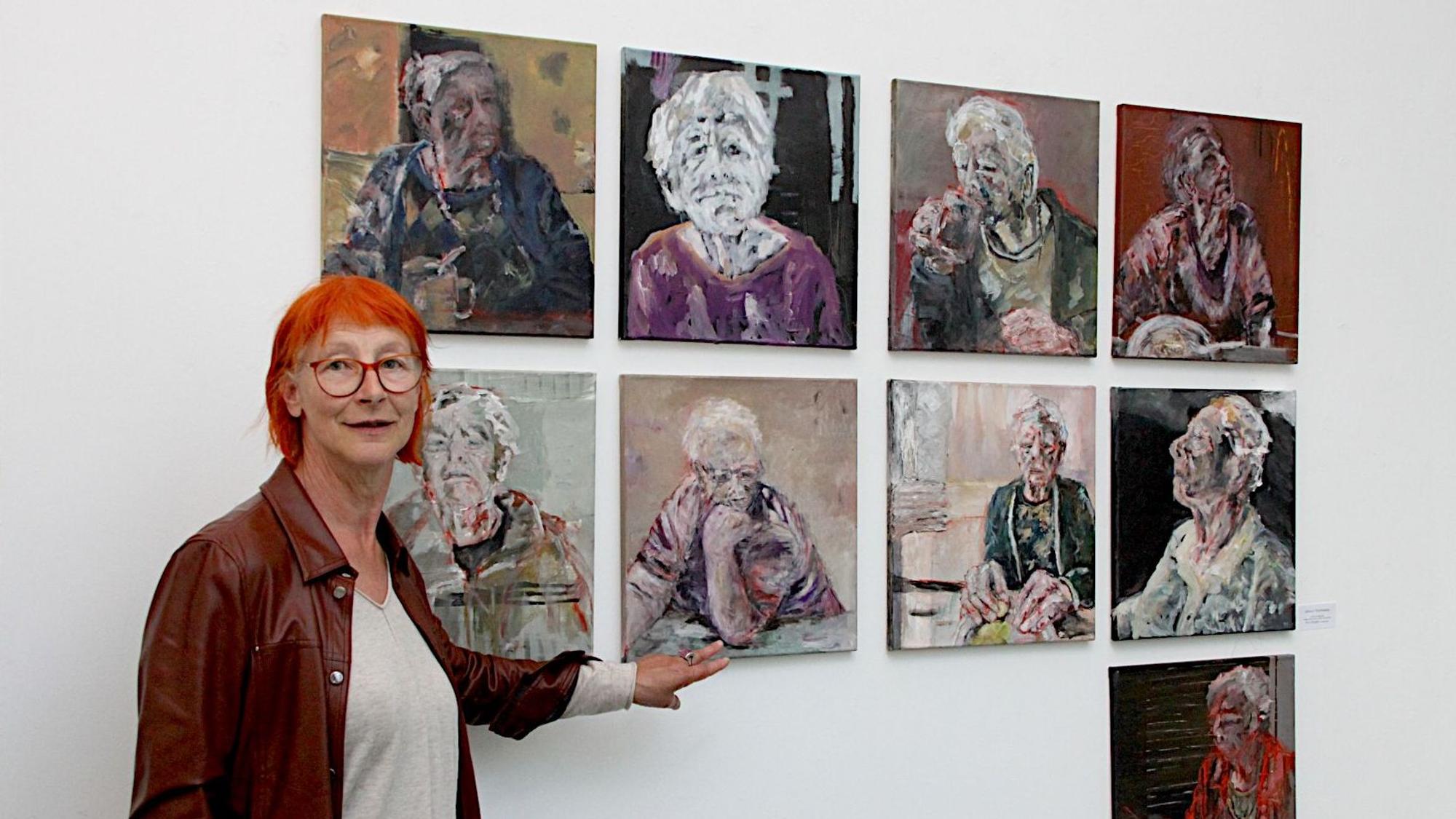 Die Künstlerin Eva Görgen, die rote Haare und eine Lederjacke trägt, steht vor neun ihrer Werke, die jeweils ein Porträt einer älteren Frau zeigen.