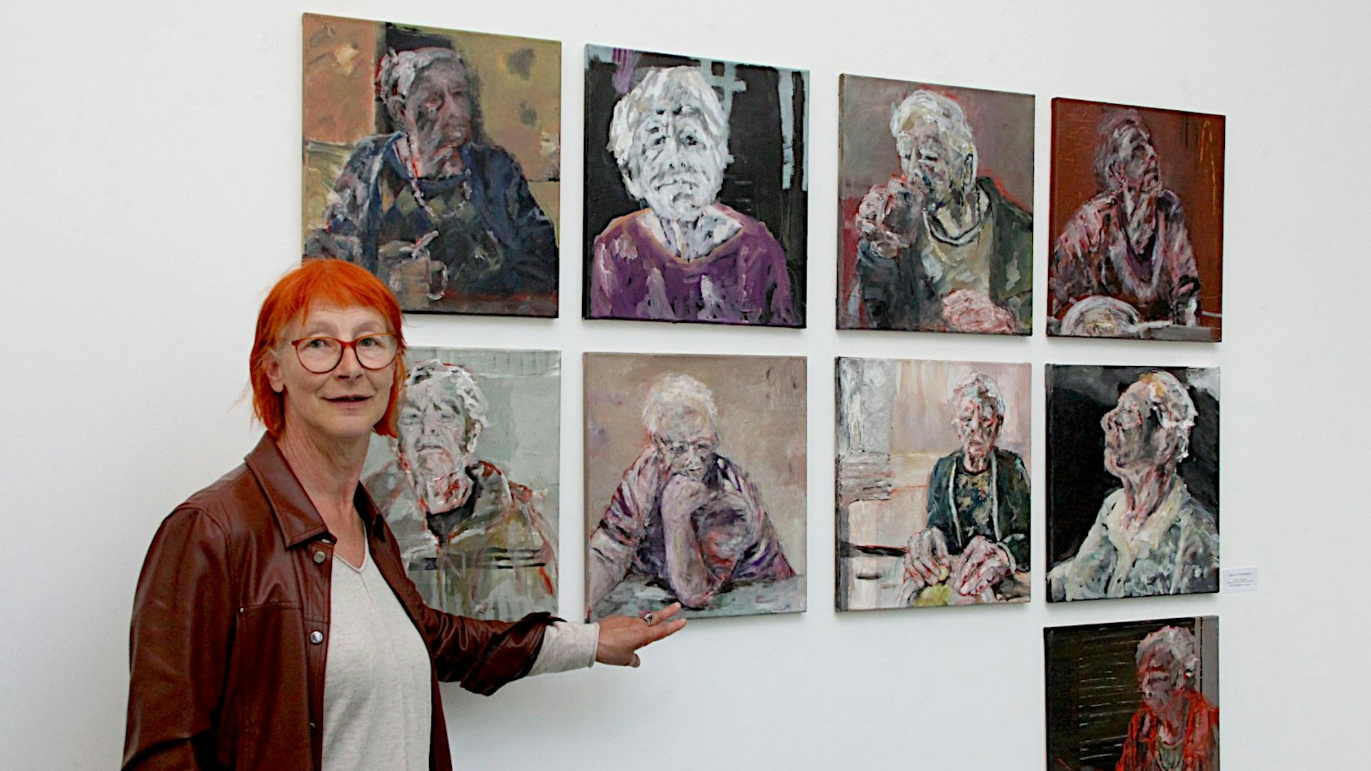 Die Künstlerin Eva Görgen, die rote Haare und eine Lederjacke trägt, steht vor neun ihrer Werke, die jeweils ein Porträt einer älteren Frau zeigen.