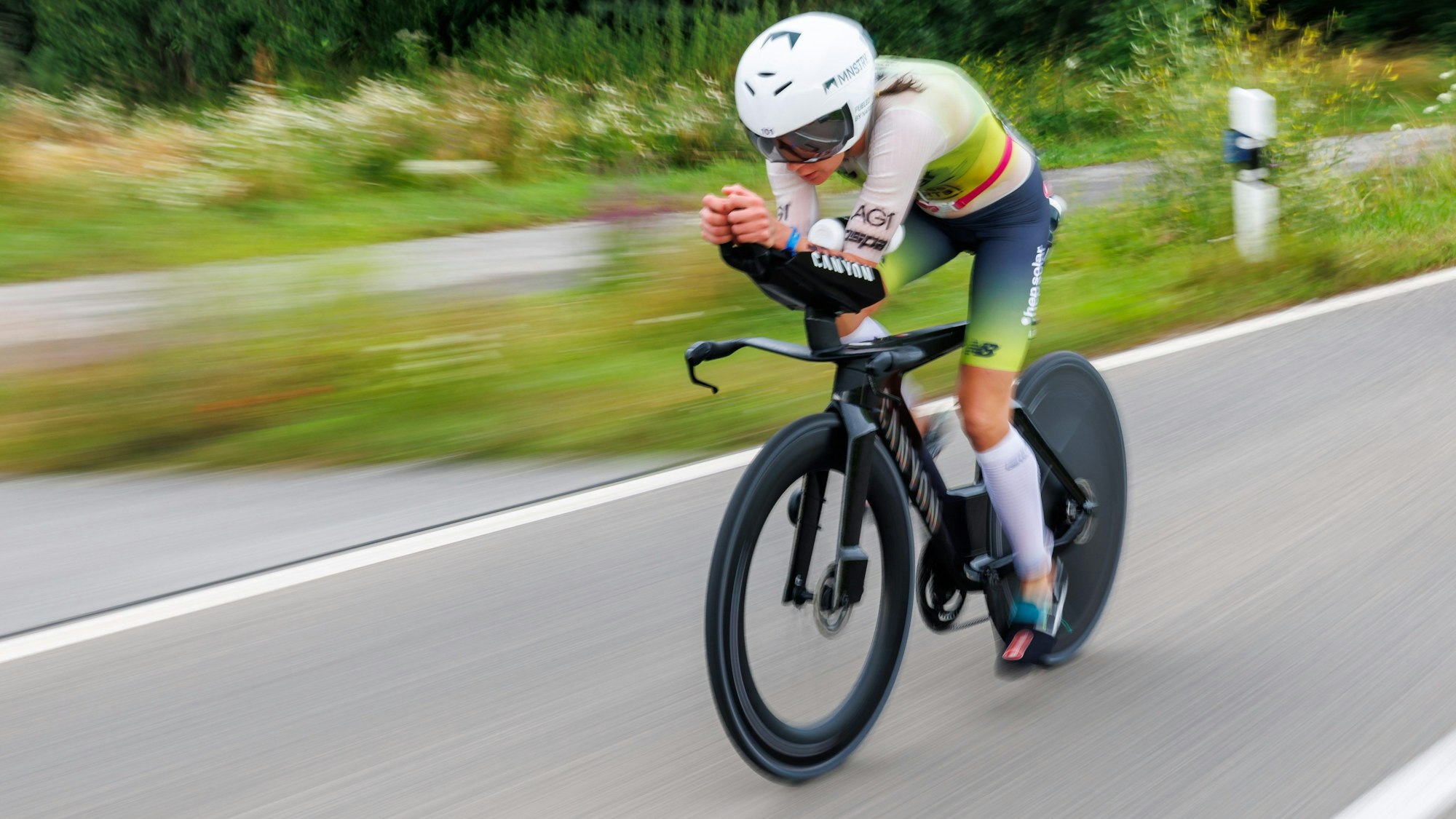 Eine Person fährt auf einem Fahrrad über eine asphaltierte Straße. Im Hintergrund sind verschwommenWiesen und Blumen zu sehen.