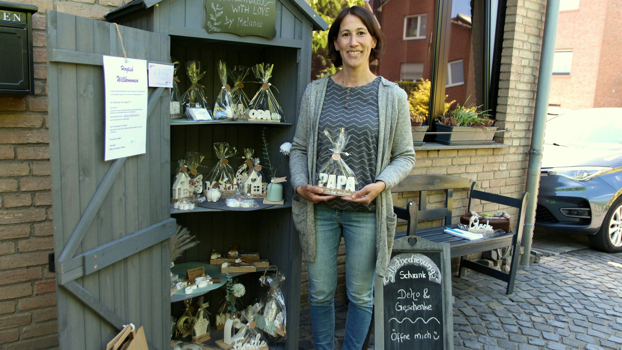 Eine dunkelhaarige Frau steht von einem Holzhäuschen, in dem Keramikarbeiten und andere Geschenkideen zu finden sind.