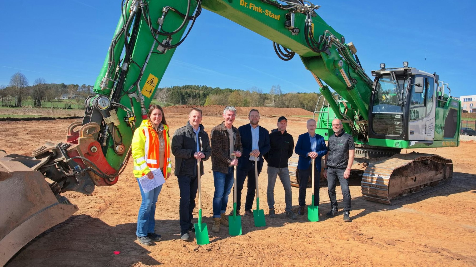 Spatenstich im neuen Mechernicher Baugebiet „Auf der Wäsche“: (v. l.) Maren Müsch, Mario Dittmann, Werner Regh, Thomas Hambach, Sven Gralka, Dr. Hans-Peter Schick und Hans Spirk, dahinter ein grüner Raupenbagger.