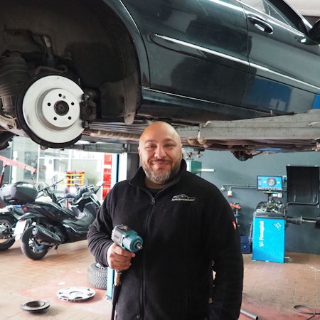 Das Bild zeigt Mhedi Ghaffari in seiner Werkstatt, hinter ihm mehrere Motorräder und ein Auto auf einer Hebebühne. In der Hand hält er einen Schlagschrauber.