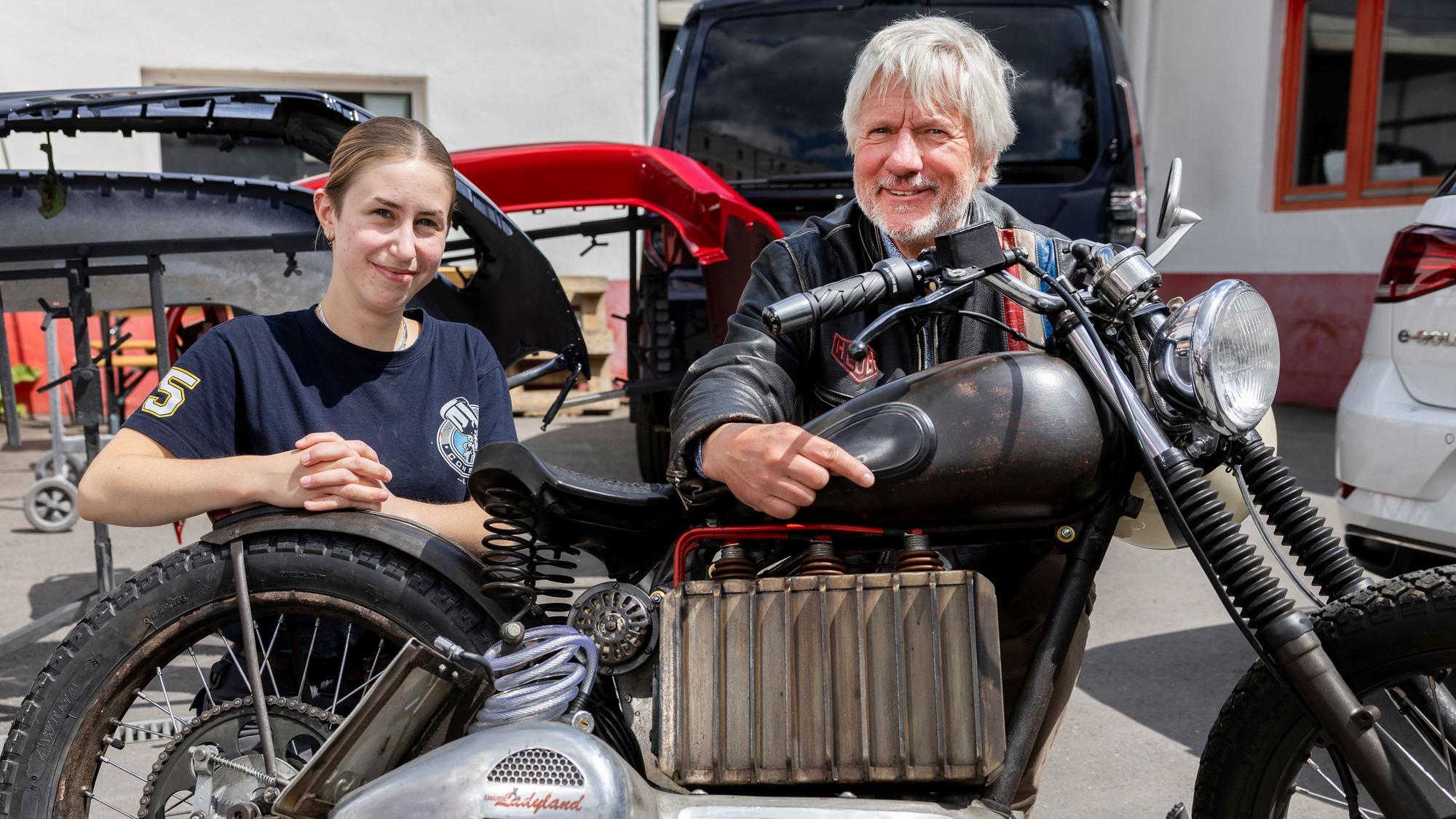 Eine junge Frau und ein Mann mit Bart hocken hinter einem Motorrad.