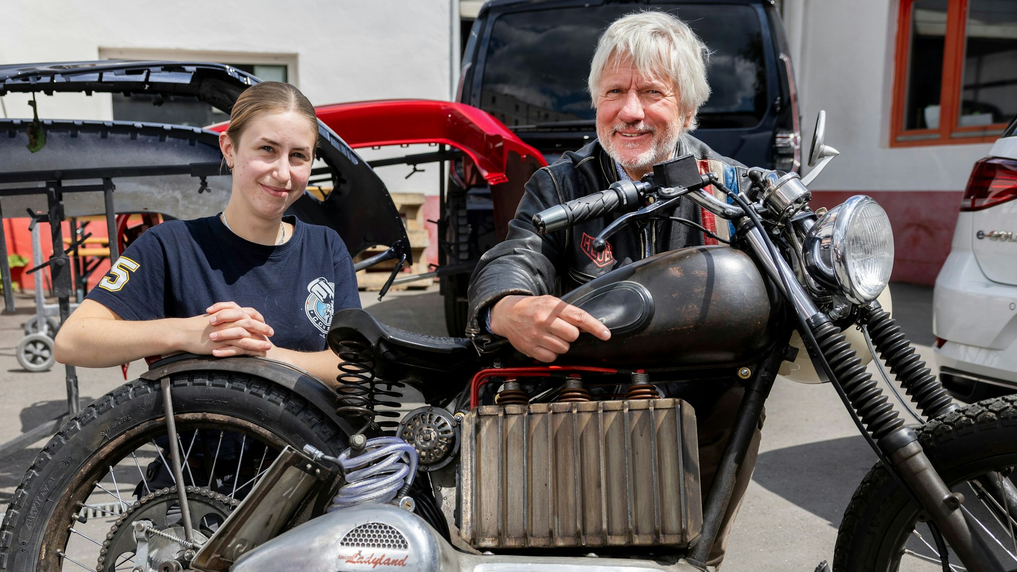 Eine junge Frau und ein Mann mit Bart hocken hinter einem Motorrad.