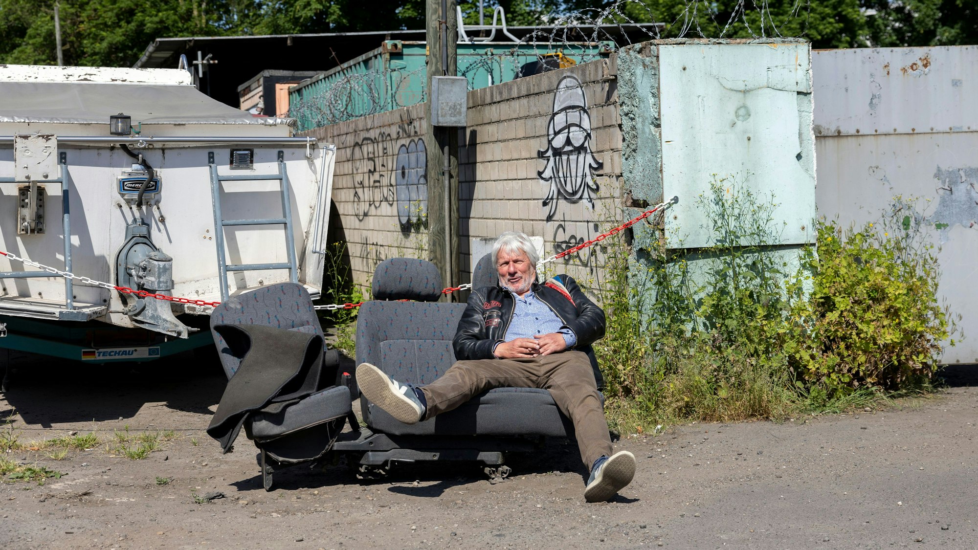 Auf einer ausgebauten Autositzbank, die am Straßenrand steht, sitzt ein Mann mit ausgestreckten Beinen.