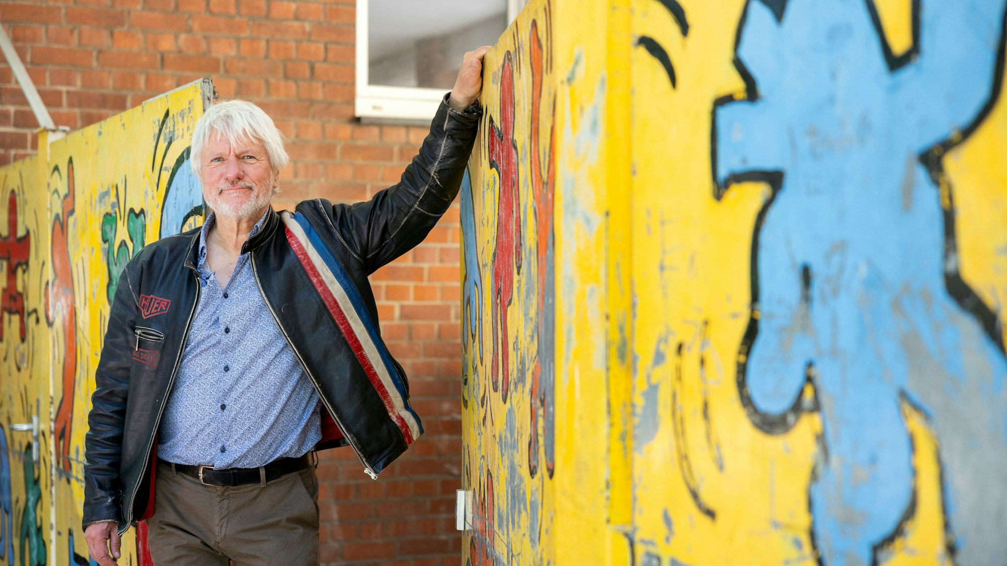 Ein Mann in Lederjacke und weißem Haar hält ein buntbemaltes Tor auf.