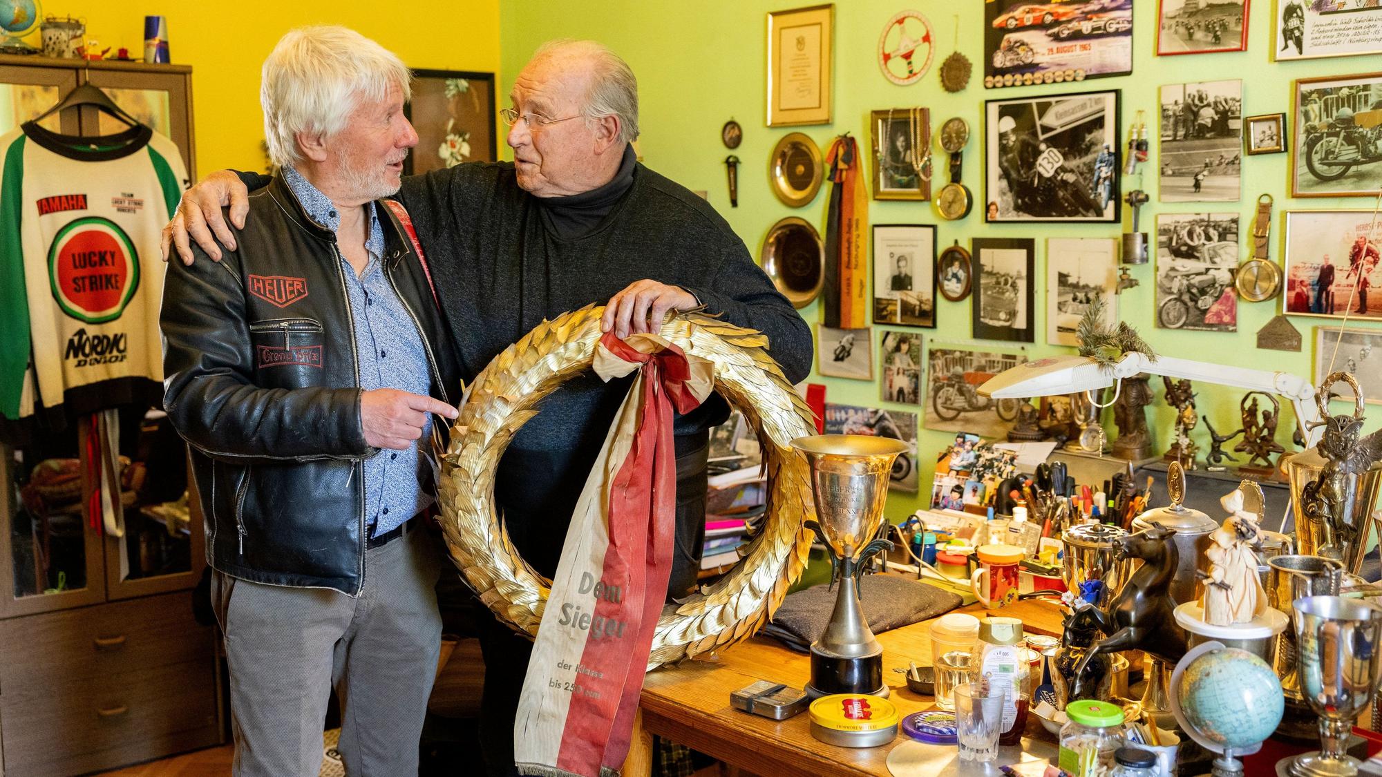 Ein Mann mit weißem Haar und ein älterer, der einen goldenen Siegerkranz hält, stehen in einem Raum, der voll ist mit Pokalen und Rennfahrer-Memorabilia.
