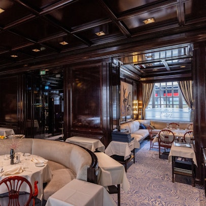 Der Restaurantraum ist zu sehen ohne Gäste. Hell bespanntes Holzmobiliar, dunkel getäfelte Wände und Decke.