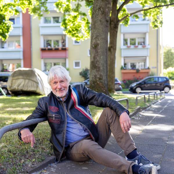 Ein Mann sitzt auf dem Bürgersteig am Rande einer von Metall-Absperrungen begrenzten Grünfläche.