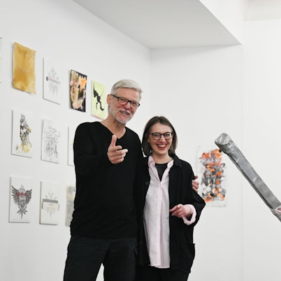 Wolfgang Voigt und Valerie Knoll posieren lachend. Voigt gestikuliert für die Kamera. Hinter ihnen sind von verschiedenen Künstlern gestaltete Variationen des Kompakt-Doppeladlers zu sehen. Neben ihnen eine Baumskulptur von Albert Oehlen.