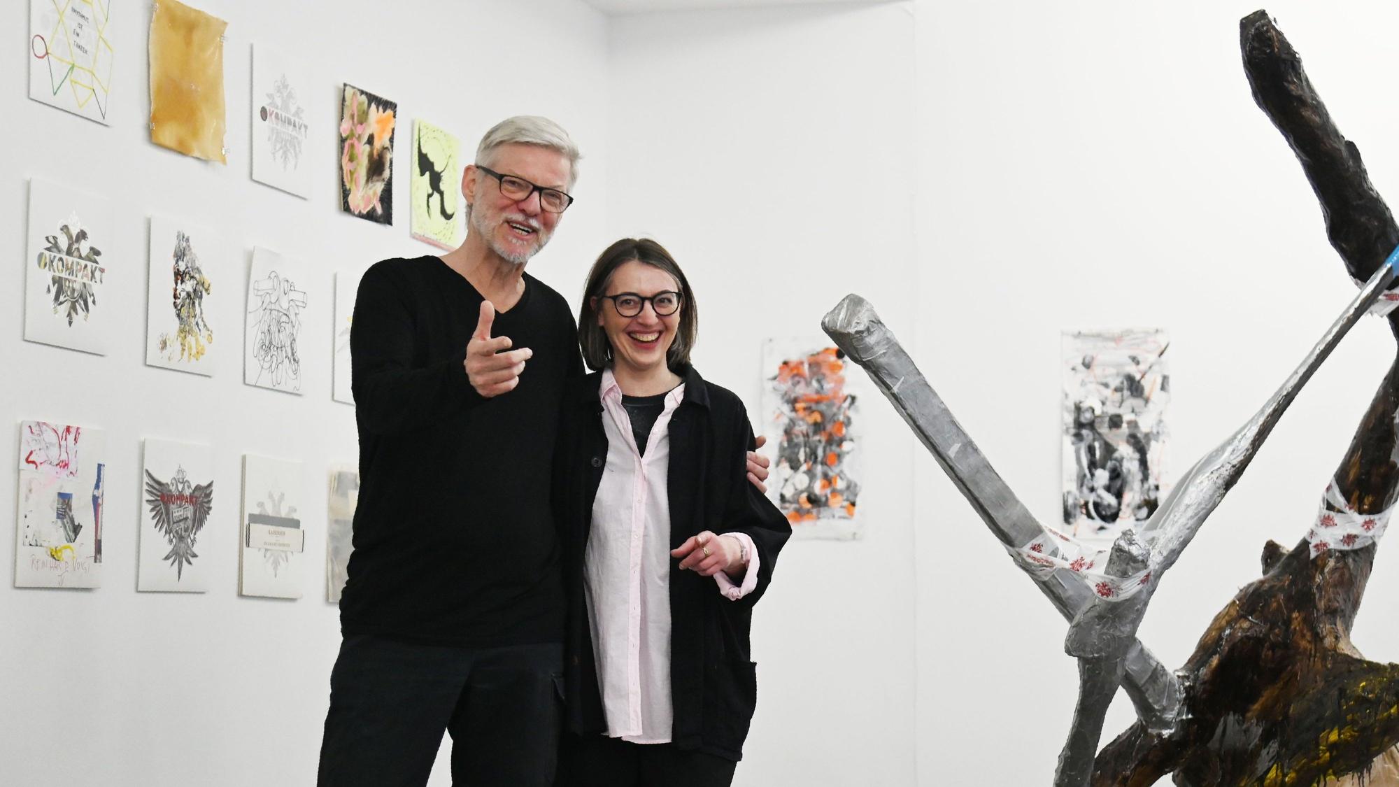 Wolfgang Voigt und Valerie Knoll posieren lachend. Voigt gestikuliert für die Kamera. Hinter ihnen sind von verschiedenen Künstlern gestaltete Variationen des Kompakt-Doppeladlers zu sehen. Neben ihnen eine Baumskulptur von Albert Oehlen.
