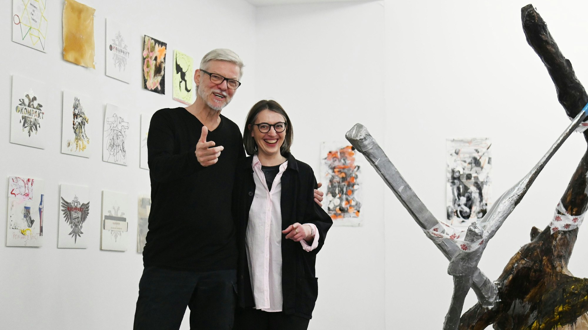 Wolfgang Voigt und Valerie Knoll posieren lachend. Voigt gestikuliert für die Kamera. Hinter ihnen sind von verschiedenen Künstlern gestaltete Variationen des Kompakt-Doppeladlers zu sehen. Neben ihnen eine Baumskulptur von Albert Oehlen.