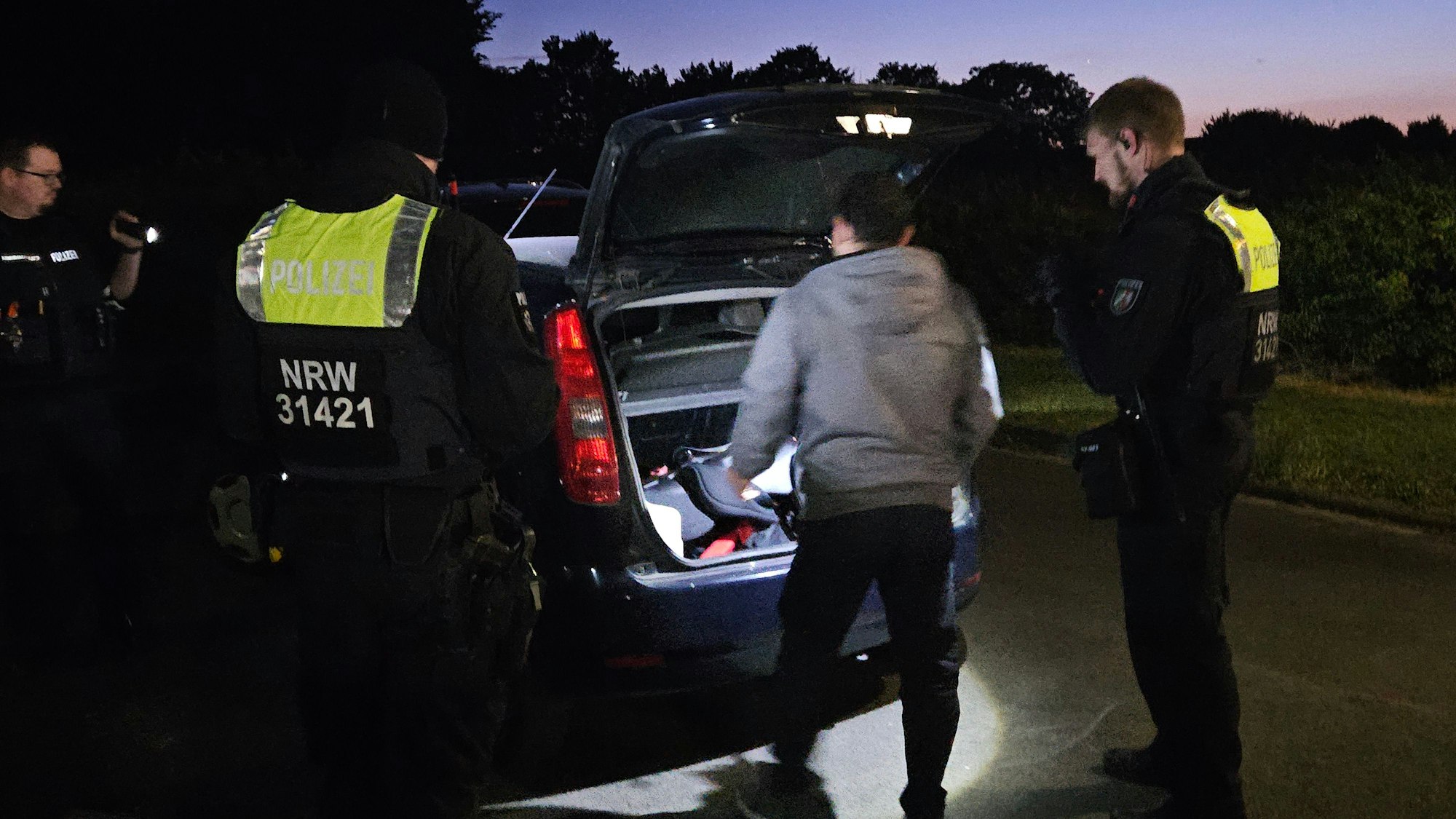Zu sehen sind Polizisten, die einen Kofferraum in Augenschein nehmen, und ein Autofahrer.