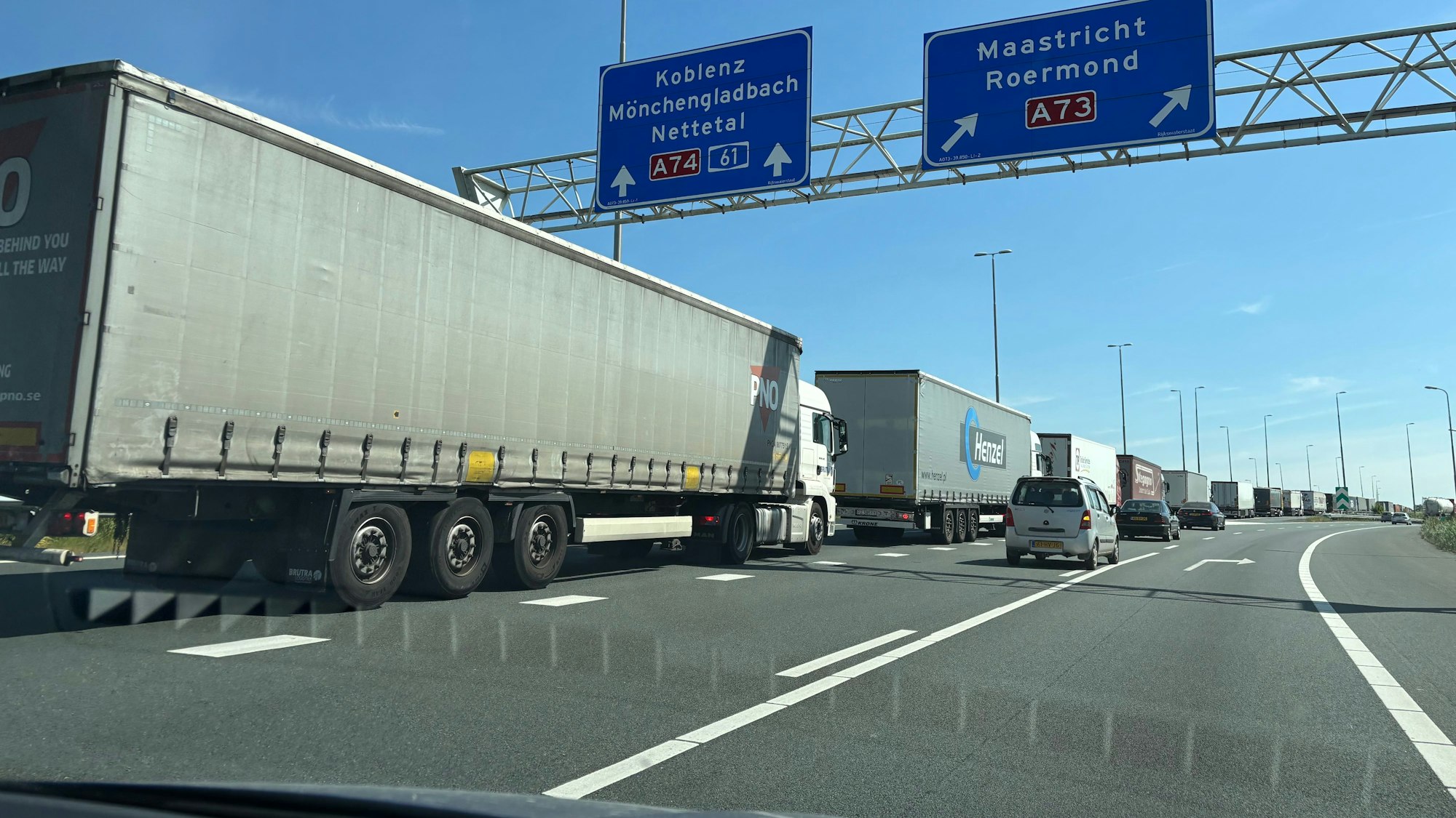 LKW stehen hintereinander im Stau. Über ihnen hängt ein Schild mit der Aufschrift „Koblenz“, darunter „Mönchengladbach“ und „Nettetal“, „A 74 61“.