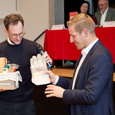 Das Bild zeigt, wie Waasem Arbeitshandschuhe mit der Aufschrift SPD an Markus Ramers übergibt. In der anderen Hand hält Waasem einen Fresskorb